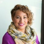 Smiling woman in a purple dress and scarf holding a tablet indoors.